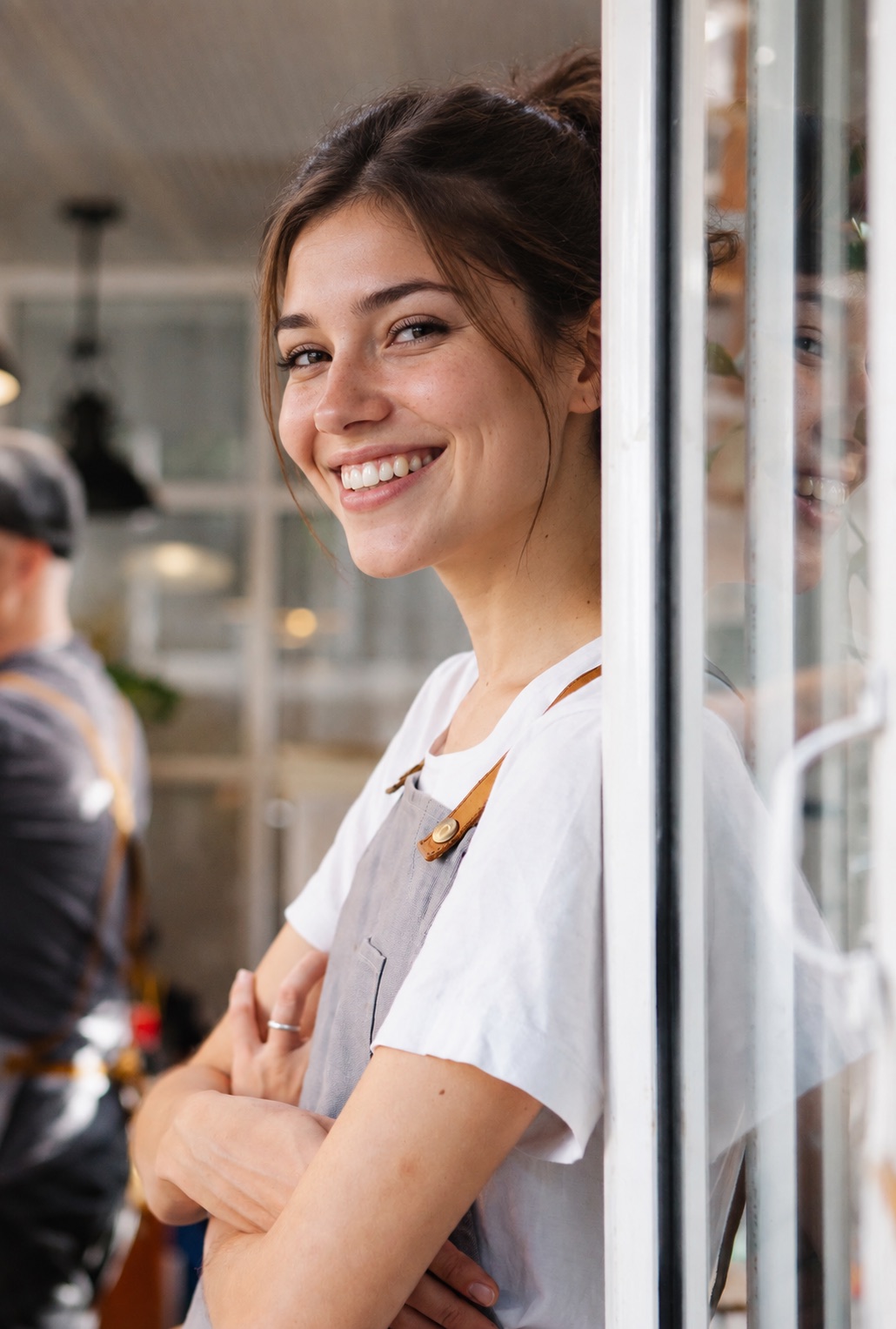 Eine junge Frau in Schürze lächelt an der Tür eines Cafés.