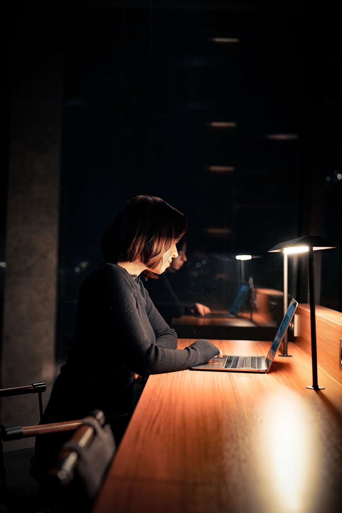 Eine Frau arbeitet abends allein am Laptop in einem dunklen Büro, nur die Schreibtischlampe brennt.