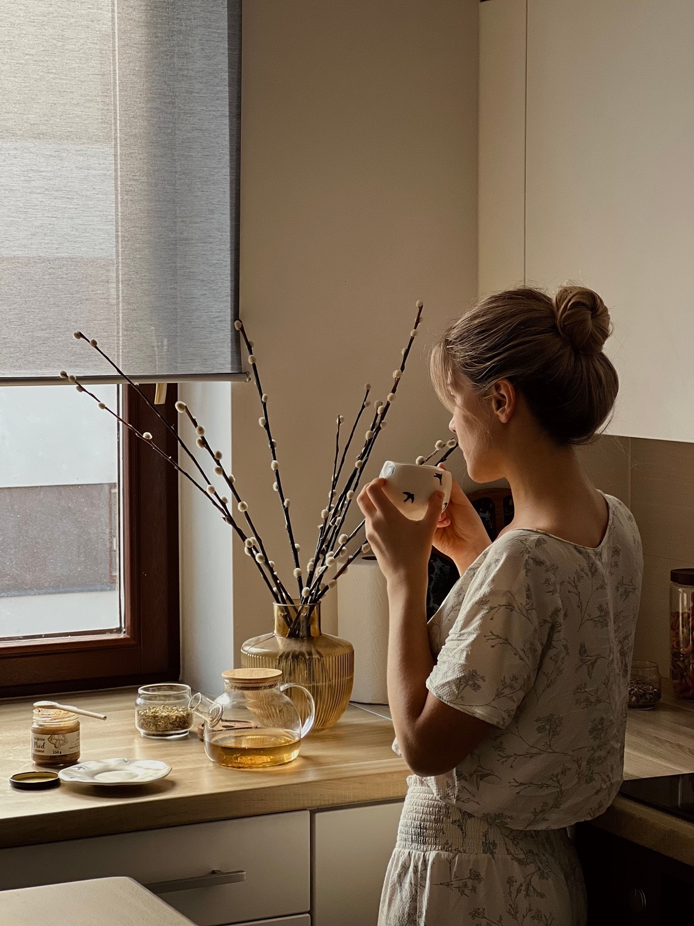 Eine Frau steht mit einer Tasse Tee am Küchenfenster im weichen Morgenlicht.
